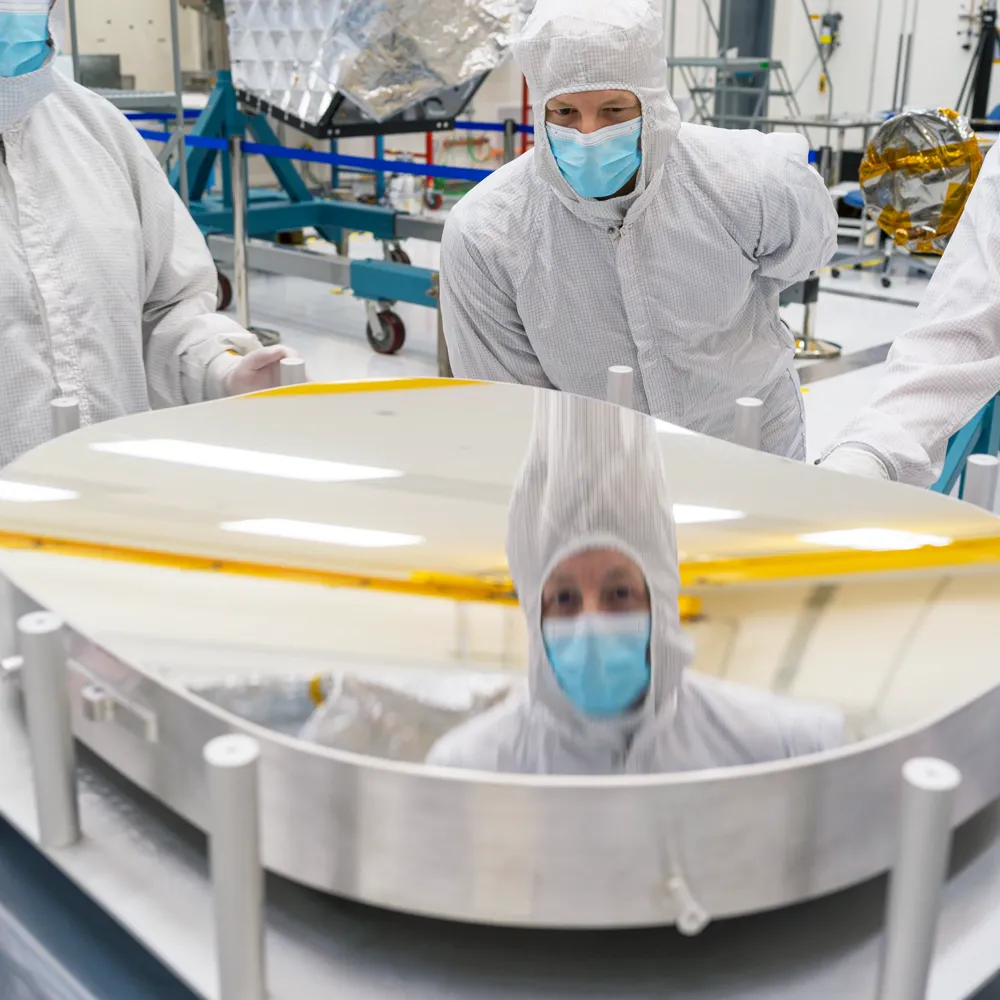 Engineers in cleanroom dress inspecting a large flight mirror